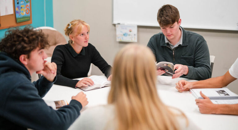 Students in a classroom setting, reading books and engaging in discussion around a table.