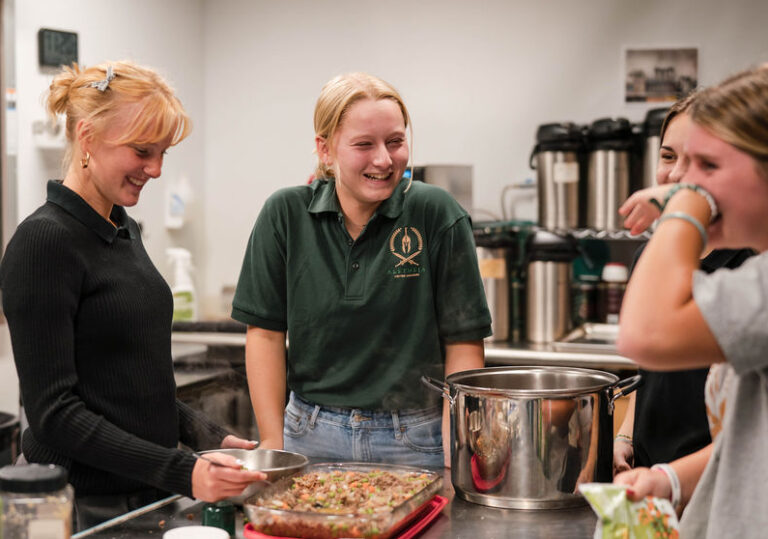 Aletheia campus tour: Students laughing while preparing food in a kitchen, pot and casserole dish visible.