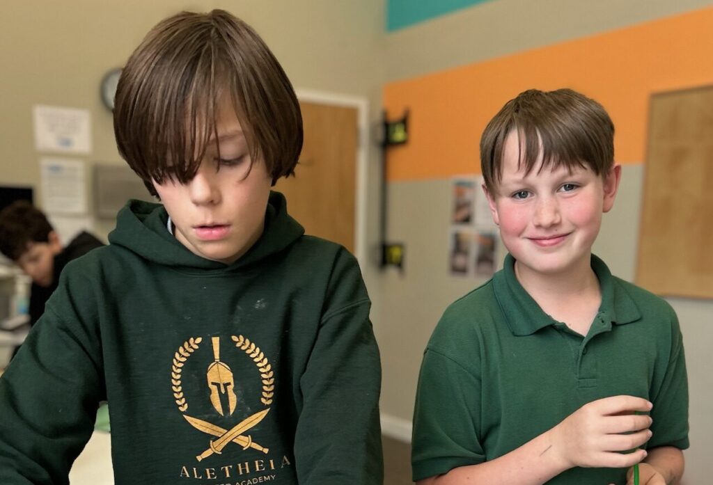 Two boys in green shirts, one wearing an Aletheia United Academy hoodie, stand indoors.
