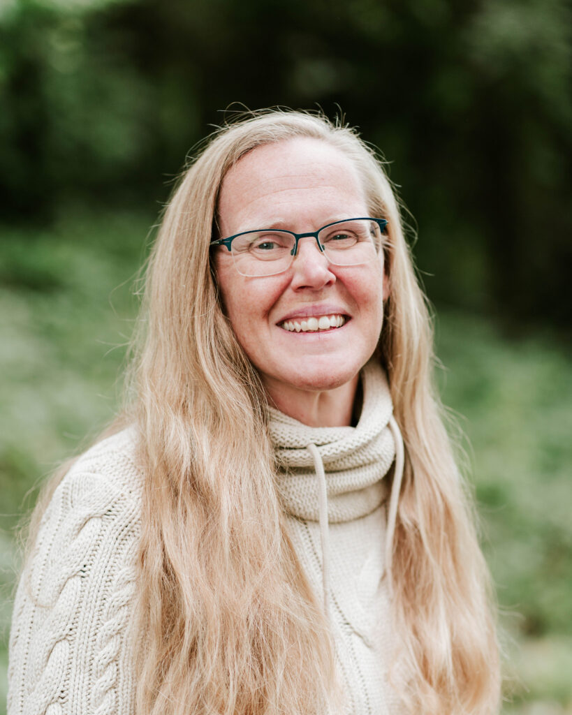 Portrait of a smiling woman with long blonde hair and glasses wearing a cream-colored sweater.