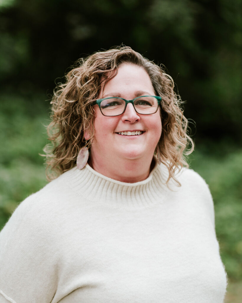 Portrait of a smiling woman with curly hair wearing glasses and a cream sweater.