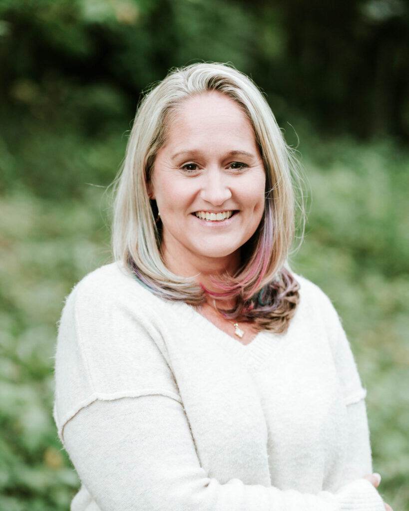 Smiling woman with blonde hair and colorful highlights wearing a white sweater.