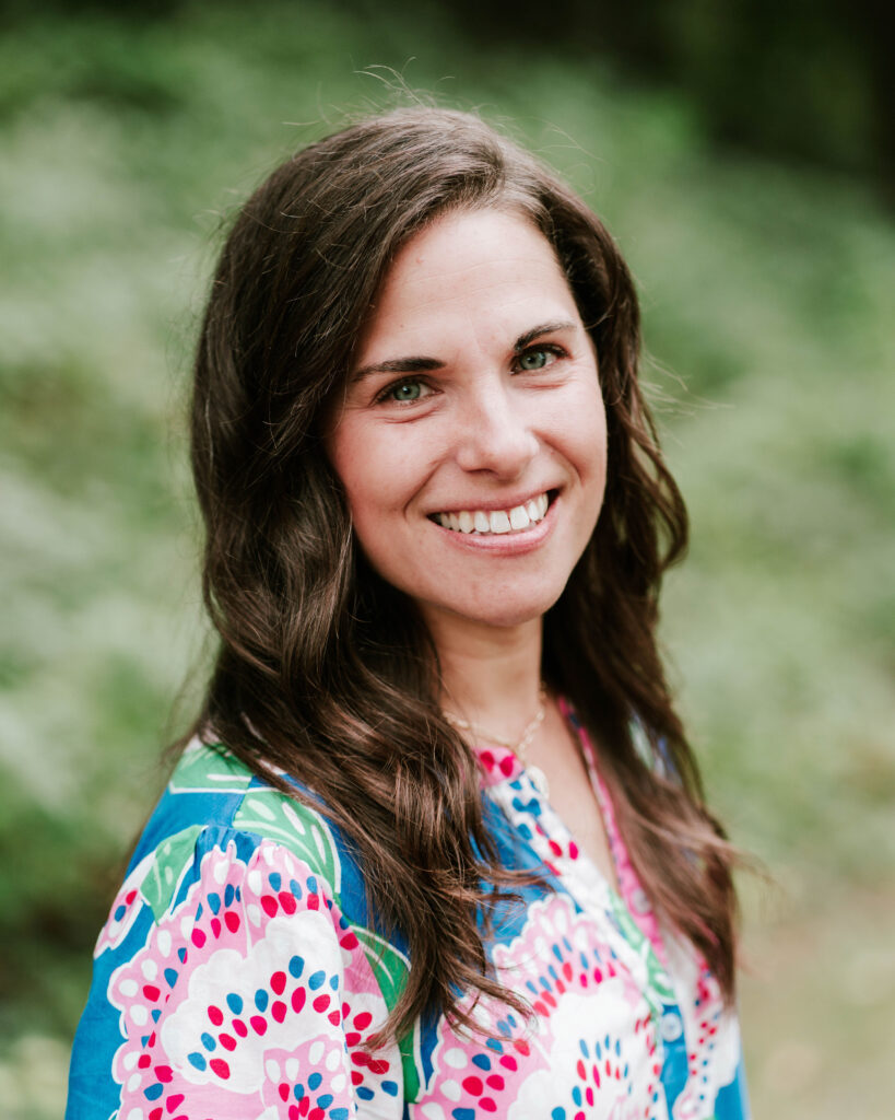 Smiling woman with long brown hair wearing a colorful patterned top.