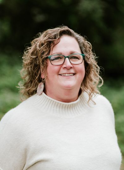 Portrait of a smiling woman with curly hair wearing glasses and a cream sweater.