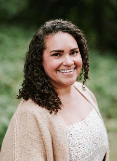Smiling woman with curly hair wearing a floral dress and cardigan. Portrait of a Graduate.