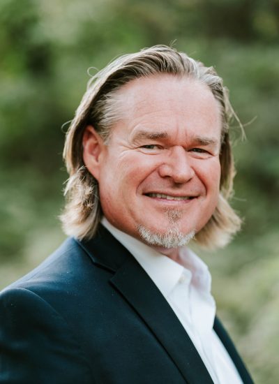 Portrait of a graduate in a suit, smiling outdoors with long blond hair.