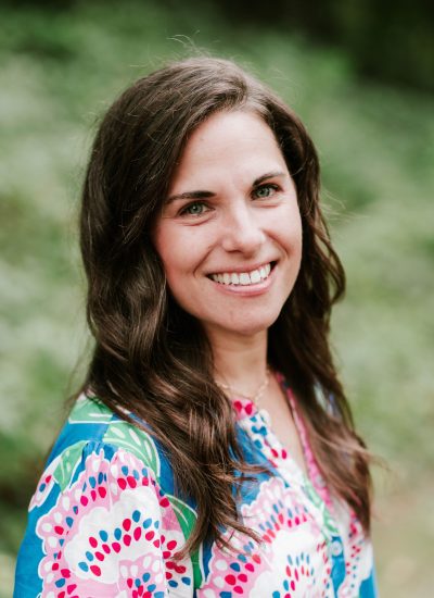 Smiling woman with long brown hair wearing a colorful patterned top.